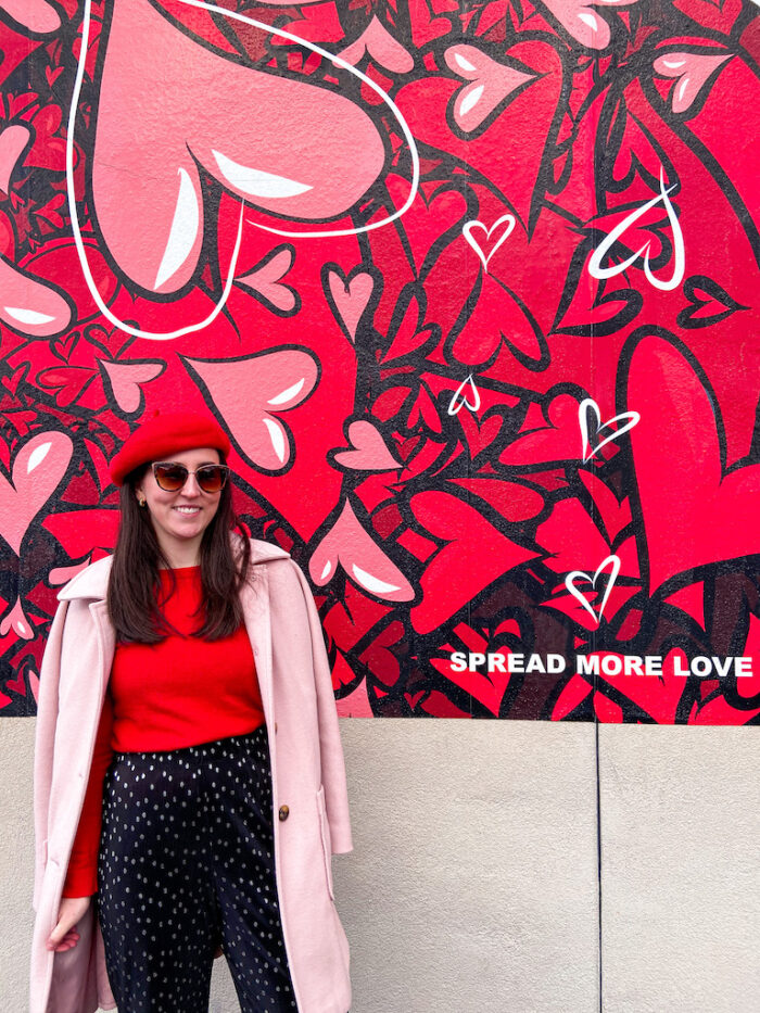 Spread Love This Weekend! Image description: red and pink hearts mural, red beret, red sweater, pink coat, black polka dot pants.