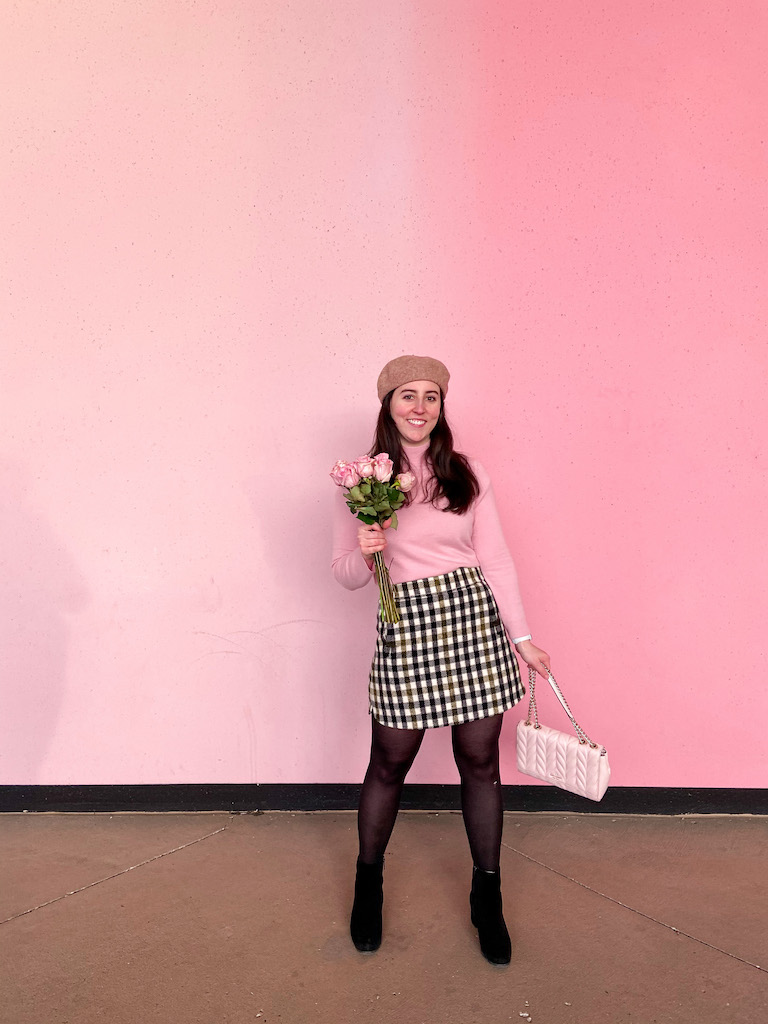 A Chic Parisian & Thrifted Look for Valentine's Day. Image description: pink sweater, gingham skirt, black tights, black suede booties, pink handbag, pink mural wall.