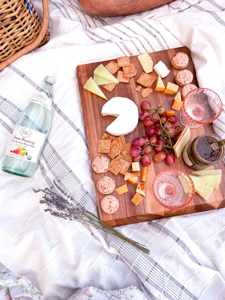 Perfect summer picnic - Image description: wooden board a top a white picnic blanket with cheese, grapes, crackers, jam, macarons, and sparkling water.