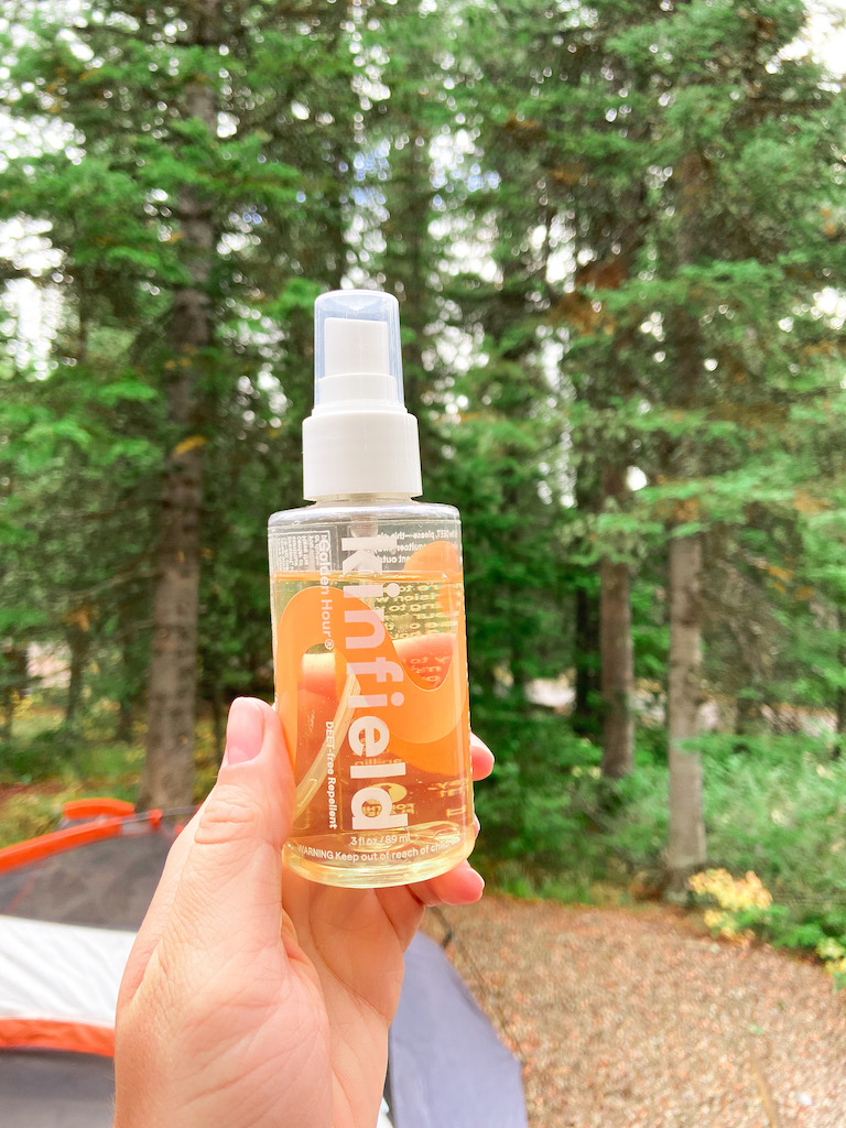 Image description: a white woman holding a bottle of Kinfield's golden hour bug spray in front of a tent and trees.