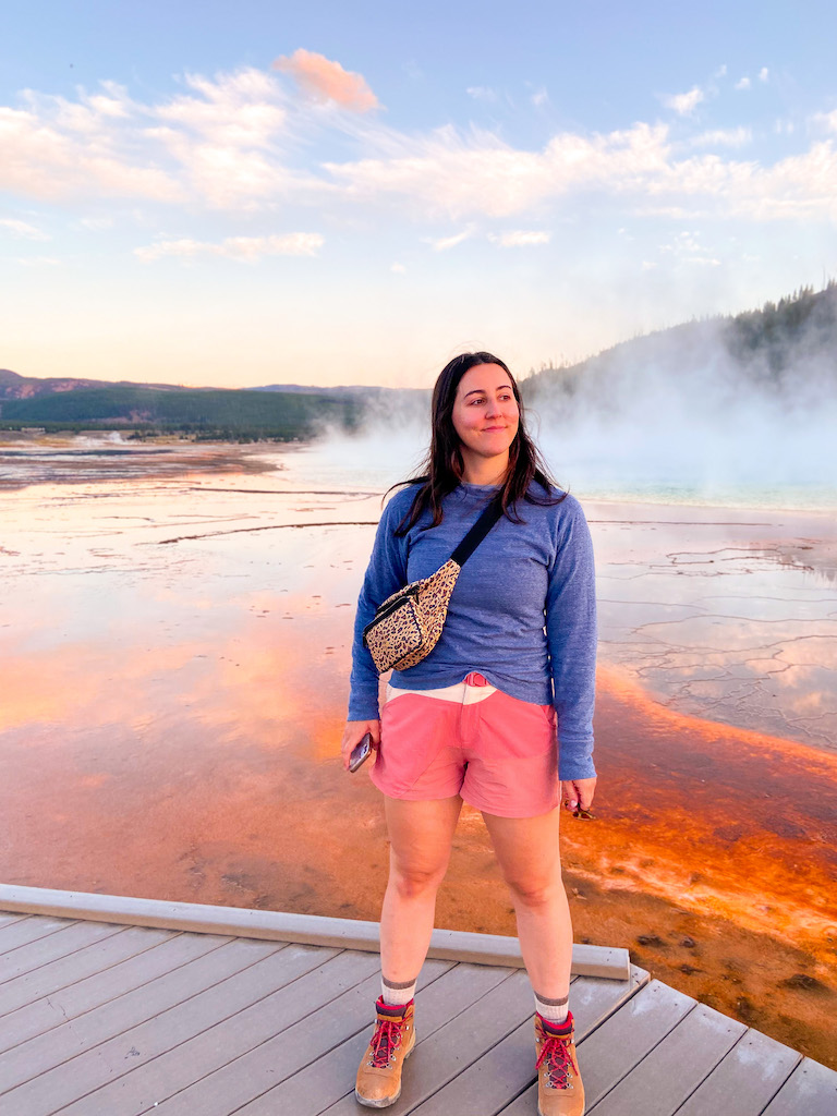 Image description: a white woman standing on the platform in front of Yellowstone National Park's Grand Prismatic Spring. She is wearing a blue pullover sweater, pink shorts, a leopard print bag, and brown hiking books. The spring in the background has steam and has red, orange and brown coloring.