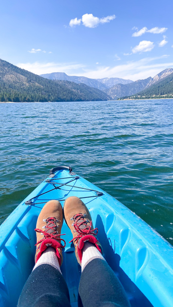 Image description: a pair of hiking boots at the front of a blue kayak. The kayak is on the water and is facing a range of mountains right outside the lake.