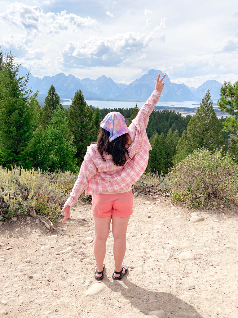 My Ultimate Packing List for a National Park Trip image description: a white woman facing away from the camera, toward the Grand Teton mountains. She has her hands up and is wearing a tie dye bandana, a pink flannel shirt, pink shorts, and black sandals.