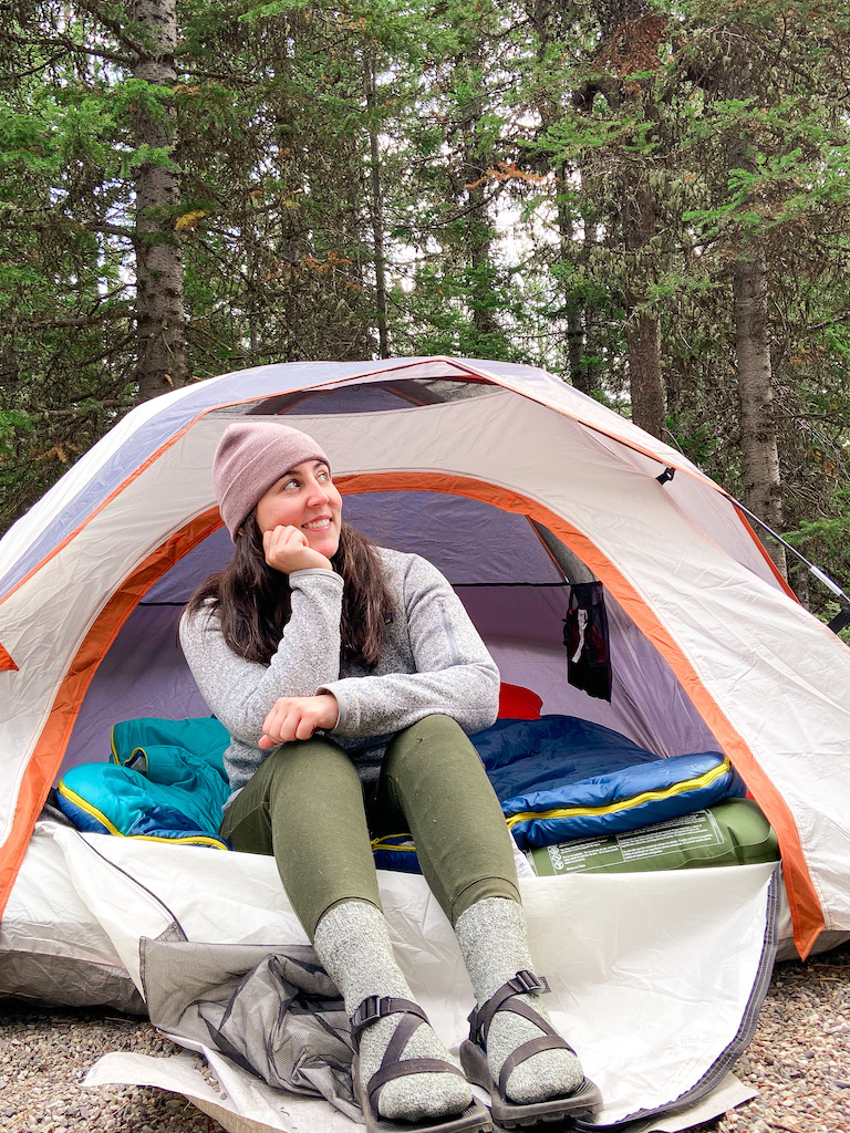 My Ultimate Packing List for a National Park Trip. Image description: a white woman sitting in the entrance of a tent in front of trees. She is wearing a beanie, gray pullover, green joggers, gray socks, and black Chaco sandals. There is a sleeping bag atop a sleeping bag in the tent behind her.