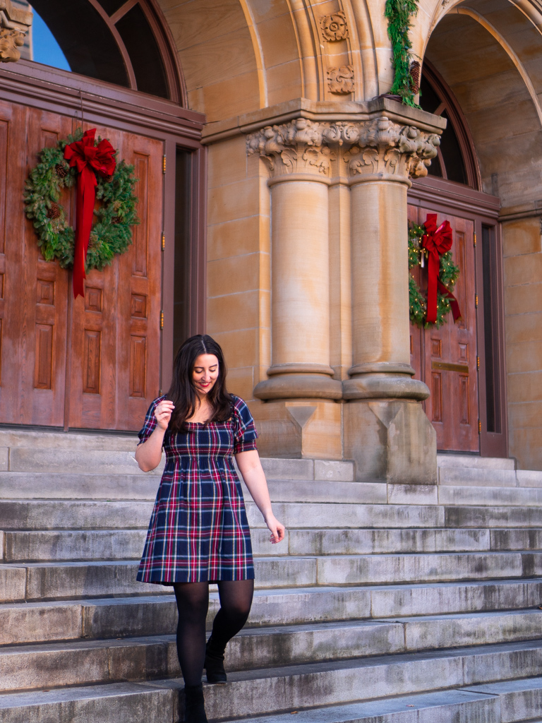 How to wear a tartan nap dress for Christmas. Image description: a white woman standing on stairs in front of a Christmas wreath hung on a door. She is wearing a navy tartan dress, black tights, and black ankle boots.