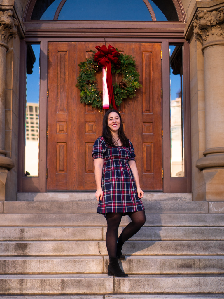 How to wear a tartan nap dress for Christmas. Image description: a white woman standing on stairs in front of a Christmas wreath hung on a door. She is wearing a navy tartan dress, black tights, and black ankle boots.