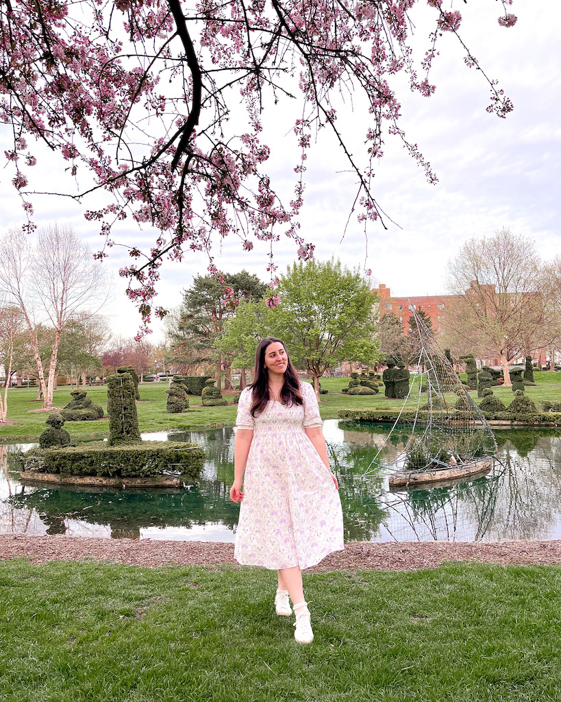 5.21.22 weekend reading - Image description: a white woman is standing in a park with green hedges behind her. She is wearing a white midi length dress and white sneakers. A cherry blossom tree hangs above her in the middle of the photo.